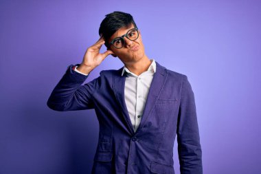 Ceket ve gözlük takan yakışıklı genç iş adamı, mor arka planda endişeli ve stresli. Alnında bir sorun var diye endişeli ve bunalımda.