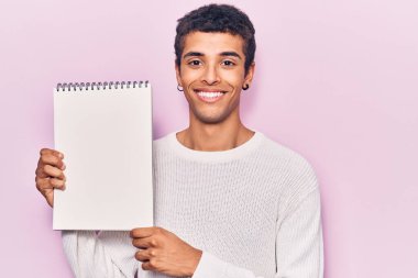 Genç Afrikalı Amerikalı bir adam elinde defterle mutlu ve pozitif görünüyor. Kendine güvenen bir gülümsemeyle dişlerini gösteriyor. 