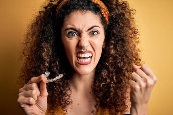 Young beautiful woman with curly hair and piercing holding dental aligner orthodontic annoyed and frustrated shouting with anger, crazy and yelling with raised hand, anger concept