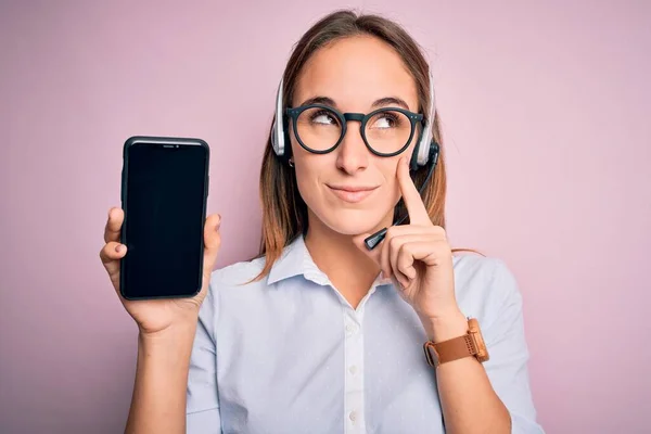 Beautiful call center agent woman working using headset holding smartphone holding screen serious face thinking about question, very confused idea