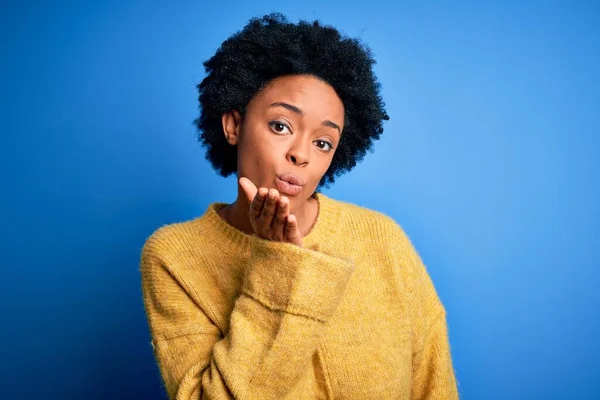 Young beautiful African American afro woman with curly hair wearing yellow casual sweater looking at the camera blowing a kiss with hand on air being lovely and sexy. Love expression.