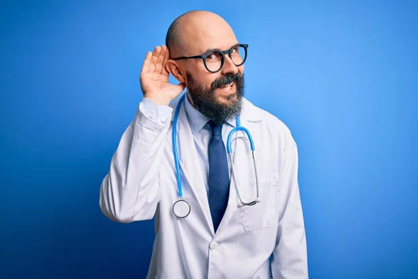 英俊的秃头医生 留着胡子 戴着眼镜 带着听诊器 带着蓝色背景微笑 两手相对地听着谣言或流言蜚语 失聪的概念 — 图库照片