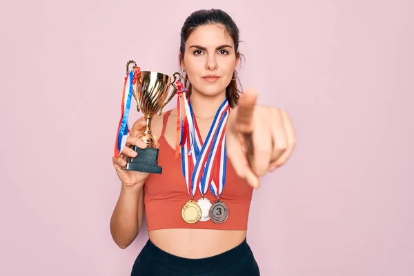 Young beautiful fitness winner athlete woman wearing sport medals and trophy pointing with finger to the camera and to you, hand sign, positive and confident gesture from the front
