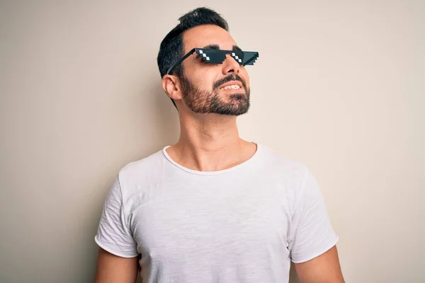 Young handsome man with beard wearing funny thug life sunglasses over white background looking away to side with smile on face, natural expression. Laughing confident.