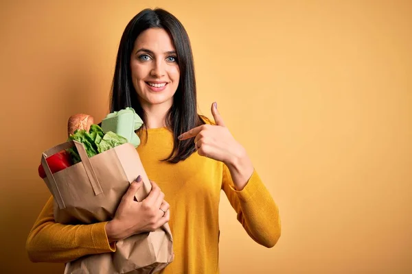 年轻的黑发女人 蓝眼睛 手里拿着购物袋里的健康食品 满脸惊讶 手指指向自己 — 图库照片