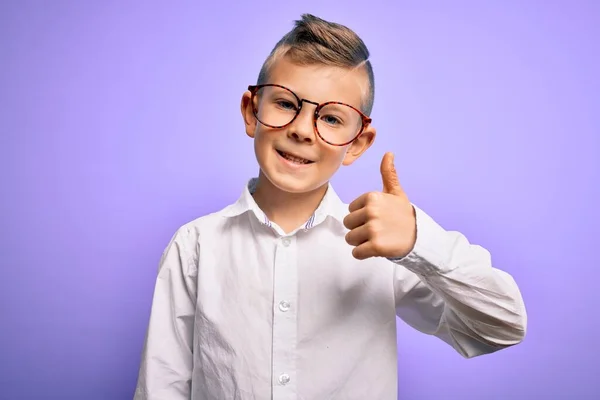 青い目の若い白人の子供は 紫色の背景に眼鏡と白いシャツを着て 手で幸せな親指を立ててジェスチャーをします 成功を示すカメラを見て表現を承認する — ストック写真