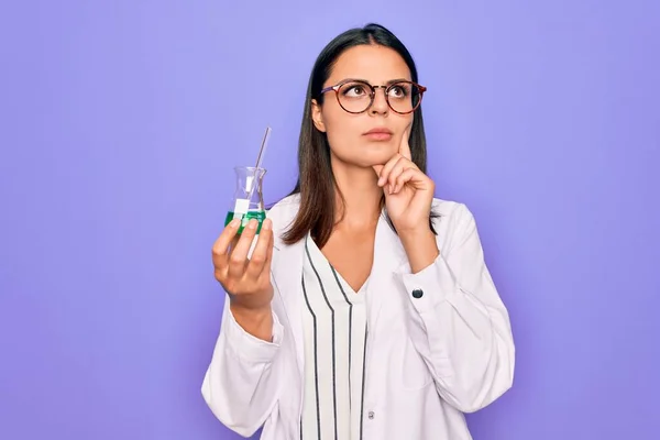 Young beautiful brunette scientist woman wearing coat and glasses holding test tube serious face thinking about question with hand on chin, thoughtful about confusing idea