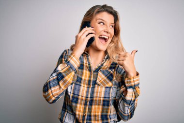 Güzel sarışın kadın beyaz arka planda akıllı telefondan konuşuyor ve gülümseyen yüzüyle baş parmağıyla onu işaret ediyor.