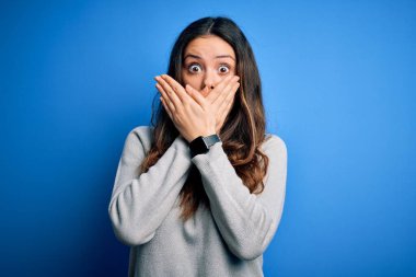 Young beautiful brunette woman wearing casual sweater standing over blue background shocked covering mouth with hands for mistake. Secret concept.