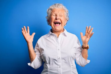 Üst düzey güzel bir kadın, zarif bir gömlek giymiş, izole edilmiş mavi arka planda durmuş, çılgın ve başarının kutlamalarını yapıyor, kollarını kaldırmış, gözlerini kapatmış, heyecanlı çığlıklar atıyor. Kazanan konsept