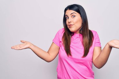 Young beautiful brunette woman wearing casual pink t-shirt standing over white background clueless and confused expression with arms and hands raised. Doubt concept. clipart