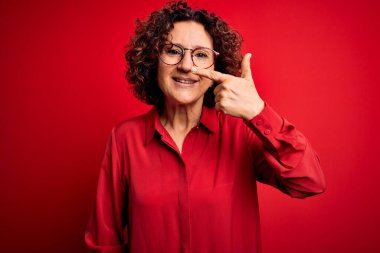 Orta yaşlı, kıvırcık saçlı, normal gömlekli ve kırmızı arka planda gözlüklü güzel bir kadın. Parmaklarıyla yüzünü ve burnunu işaret ediyor. Neşeli bir şekilde gülümsüyor. Güzellik konsepti