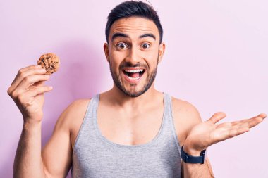 Sakallı, kolsuz tişörtlü, çikolatalı kurabiye yiyen genç yakışıklı adam zaferi mutlu bir gülümsemeyle kutluyor ve ellerini kaldırarak kazanan yüz ifadesi sergiliyor.