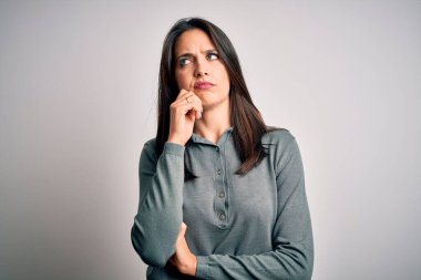 Mavi gözlü genç esmer kadın beyaz arka plan üzerine yeşil süveter giyiyor ve elleri çenesinde soru sormayı düşünüyor. Düşünceli bir ifade. Düşünceli bir yüzle gülümsüyor. Şüphe kavramı.