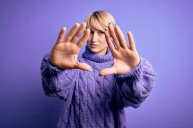 Sarışın, kısa saçlı, balıkçı yaka kazak giyen, mor arka planda elleri ve parmakları kullanarak çerçeve yapan genç bir kadın.