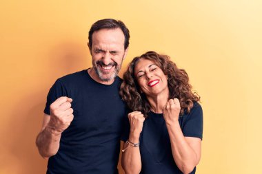 Orta yaşlı güzel bir çift, izole edilmiş sarı arka planda tişört giyip kolları havada ve gözleri kapalı bir şekilde başarıyı kutluyor.