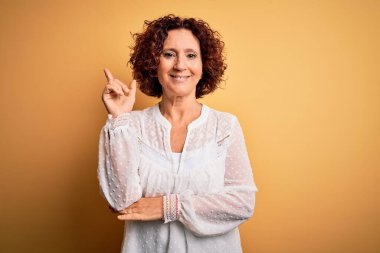 Orta yaşlı güzel kıvırcık saçlı kadın sarı arka planda rahat bir yaz elbisesi giyiyor, yüzünde kocaman bir gülümseme, eli ve parmağı kameraya bakıyor..