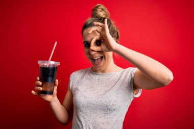 Güzel sarışın kadın kırmızı arka planda ferahlamak için kola köpüklü içecek içiyor. Gülümsüyor, el ve gözlerle parmaklarından bakıyor.