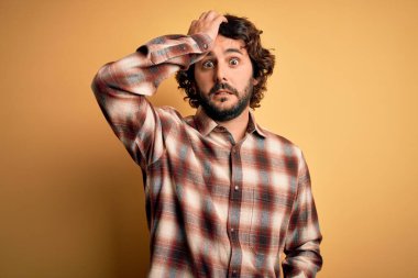 Young handsome man with beard wearing casual shirt standing over yellow background surprised with hand on head for mistake, remember error. Forgot, bad memory concept. clipart