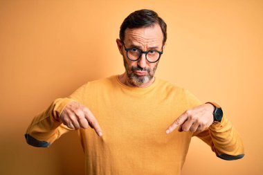 Orta yaşlı, beyaz bir adam, günlük süveter ve gözlük takıyor, sarı arka planda, üzgün ve üzgün görünüyor, parmaklarıyla yönü gösteriyor, mutsuz ve depresif..