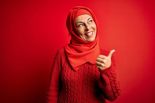 Middle age woman wearing traditional muslim hijab standing over isolated red background smiling with happy face looking and pointing to the side with thumb up.