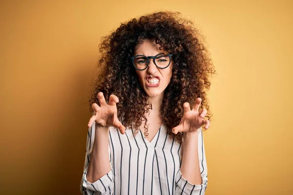 Young beautiful woman with curly hair and piercing wearing striped shirt and glasses smiling funny doing claw gesture as cat, aggressive and sexy expression