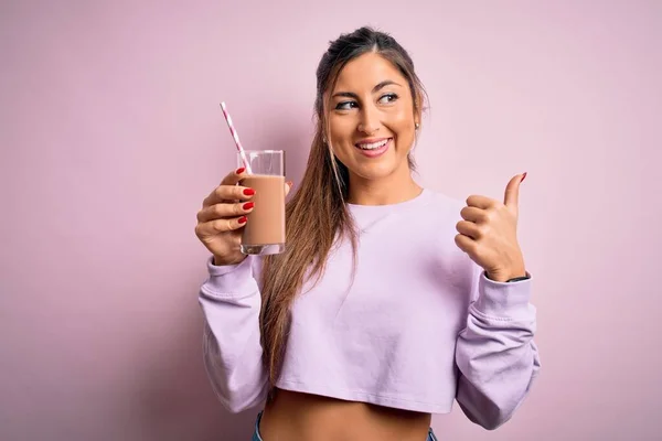 年轻美丽的健康女人在运动前喝着蛋白质巧克力奶昔 用拇指尖着身体 笑容满面的站在旁边 — 图库照片