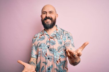 Sakallı ve dövmeli yakışıklı bir adam pembe arka planda sıradan bir çiçek gömleği giyiyor. Kolları açık gülümsüyor. Dostça karşılama, olumlu ve kendinden emin selamlar.
