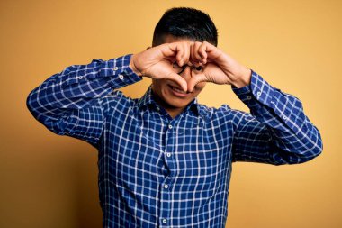 Sarı arka planda gündelik gömlek ve gözlük takan genç, yakışıklı Latin adam kalp şeklinde elleri ve parmakları ile tabelanın içinden bakıp gülümsüyor.