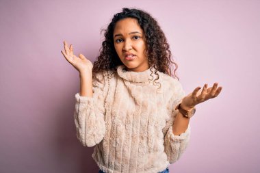 Kıvırcık saçlı, pembe arka planda dikilen ve kolları açık duran sıradan bir süveter giyen genç ve güzel bir kadın..