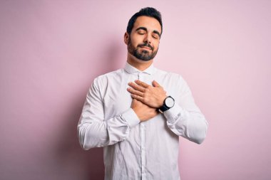 Sakallı genç yakışıklı bir adam pembe arka plan üzerinde dikiliyor elleri göğsünde, gözleri kapalı ve yüzünde minnet dolu bir jest ile gülümsüyor. Sağlık kavramı.
