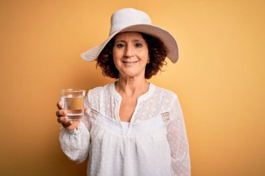 Orta yaşlı kıvırcık kadın tatilde izole edilmiş sarı arka planda bir bardak su içiyor mutlu bir yüzü var ve kendine güvenen bir gülümsemeyle dişlerini gösteriyor