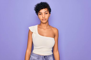 Genç ve güzel Afro-Amerikan kadın mor arka plan üzerine günlük tişört giyiyor. Yüzünde ciddi bir ifadeyle rahatlamış. Basit ve doğal bir şekilde kameraya bakıyorsun..
