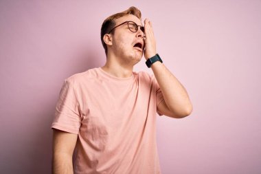 Genç, kızıl saçlı, günlük tişört giyen, izole edilmiş pembe arka planda duran, esnemekten yorgun yüzlü, göz ve ağzı elle kapatan genç bir adam. Yüzüm acıyor..