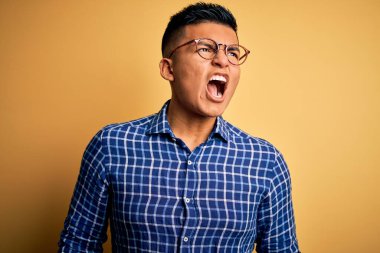 Genç, yakışıklı, latin gömlekli, sarı arka planda gözlüklü sinirli sinirli ve öfkeli bağıran öfkeyle bağıran bir adam. Öfke ve agresif kavram.
