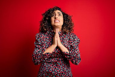 Genç ve güzel kıvırcık Arap kadın, sıradan bir çiçek elbisesi giyiyor kırmızı arka planda duruyor, elleri bağlı yalvarıyor ve dua ediyor. Yüzünde umut ifadesi var. Çok duygusal ve endişeli. Yalvarıyorum..