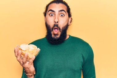 Genç Arap adam elinde patates cipsi tutuyor. Korkmuş ve ağzı açık kalmış, şaşırmış ve şaşırmış. 