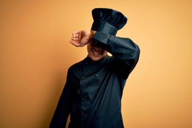 Aşçı üniforması giyen genç Brezilyalı aşçı ve izole edilmiş sarı arka planda şapkası olan neşeli ve komik kollu gözleri olan. Kör kavram.