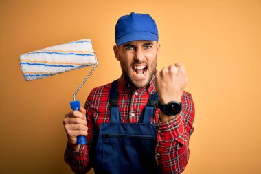 Duvarı boyayan genç yakışıklı ressam izole edilmiş sarı arka planda silindir kullanıyor. Sinirli ve sinirli bir şekilde bağırıyor, çıldırıyor ve el kaldırarak bağırıyor. Öfke kavramı.