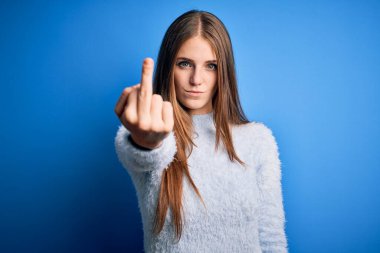 Young beautiful redhead woman wearing casual sweater over isolated blue background Showing middle finger, impolite and rude fuck off expression