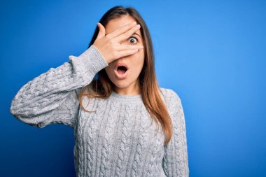 Güzel, genç bir kadın, günlük yün bir kazak giyiyor mavi izole edilmiş arka planda dikiliyor. Şok olmuş bir şekilde yüzünü ve gözlerini kapıyor, utanmış bir ifadeyle parmaklarının arasından bakıyor..