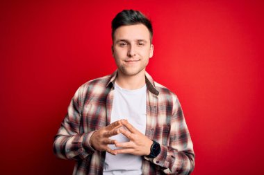 Kırmızı izole edilmiş arka plan üzerinde gündelik modern gömlekler giyen yakışıklı beyaz adam ellerini ve parmaklarını birleştirerek gülümseyerek gevşemiş ve neşeli olmasını diliyordu. Başarı ve iyimser