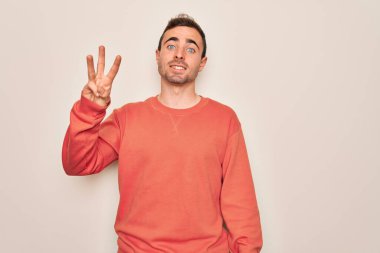Mavi gözlü genç adam beyaz arka planda dikilip üç numarayı gösterirken kendinden emin ve mutlu bir şekilde gülümsüyor..