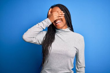 Genç Afro-Amerikalı kadın mavi izole edilmiş arka planda balıkçı yaka kazak giyerek ayakta duruyor. Gülümsüyor ve yüzünde şaşkınlıktan gözleri kapatarak gülüyor. Kör kavram.