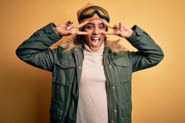 Genç Afrikalı Afro-Amerikan kayakçı kız kar sporu kıyafetleri ve kayak gözlüğü takıyor. Barış sembolü yapıyor, parmaklarının üzerinde gülümseyerek zaferi gösteriyor.