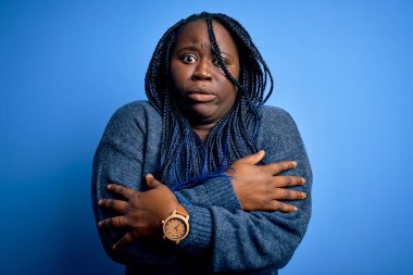 Afro-amerikan kadın örgü örüyor mavi arka planda günlük süveter giyiyor soğuk yüzünden titriyor ve donuyor yüzünde üzgün ve şok ifadesi var.