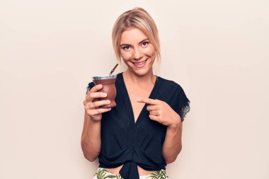 Genç, güzel, sarışın bir kadın beyaz arka planda bir fincan kahve içerken mutlu bir şekilde el ve parmağı işaret ediyor.