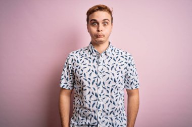 Genç, yakışıklı, yaz gömleği giyen, pembe arka planda duran, komik yüzlü kızıl saçlı adam. Havayla şişmiş ağız, çılgın bir ifade..