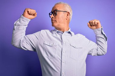 Mor arka planda çizgili gömlek ve gözlük takan orta yaşlı, beyaz bir adam gururla gülümseyen kol kaslarını gösteriyor. Fitness konsepti.