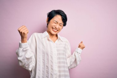 Genç, güzel Asyalı bir kız pembe arka planda tek başına dikilmiş mutlu ve heyecanlı bir şekilde kollarını kaldırarak, gülümseyerek ve başarı çığlıkları atarak zafer jesti yapıyor. Kutlama konsepti.